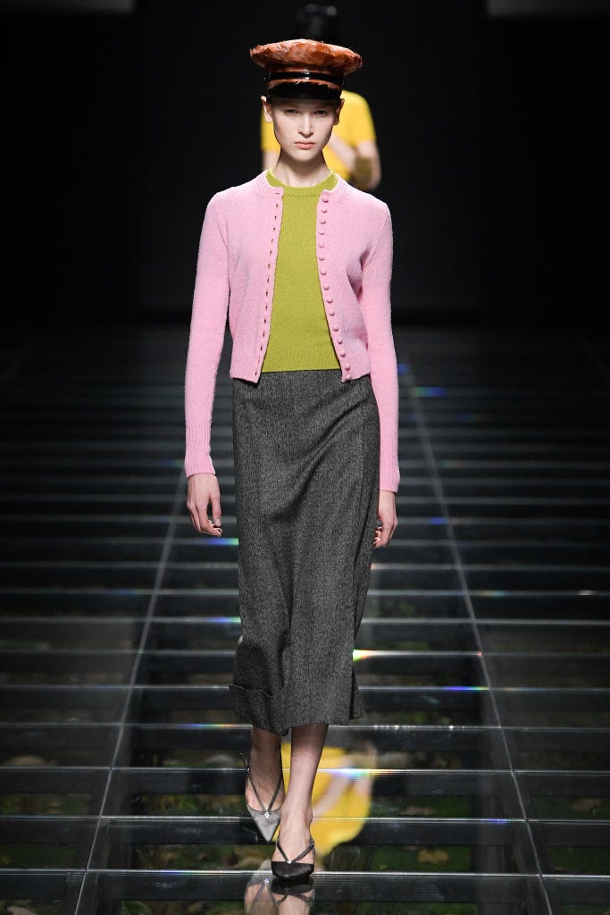 un mannequin portant un cardigan rose, un haut en tricot vert, une jupe crayon grise et des escarpins à bride arrière à bout pointu gris clair avec une casquette paperboy en cuir marron sur le défilé prada rtw automne 2024 dans un tour d'horizon des meilleures tenues de cardigan 2024