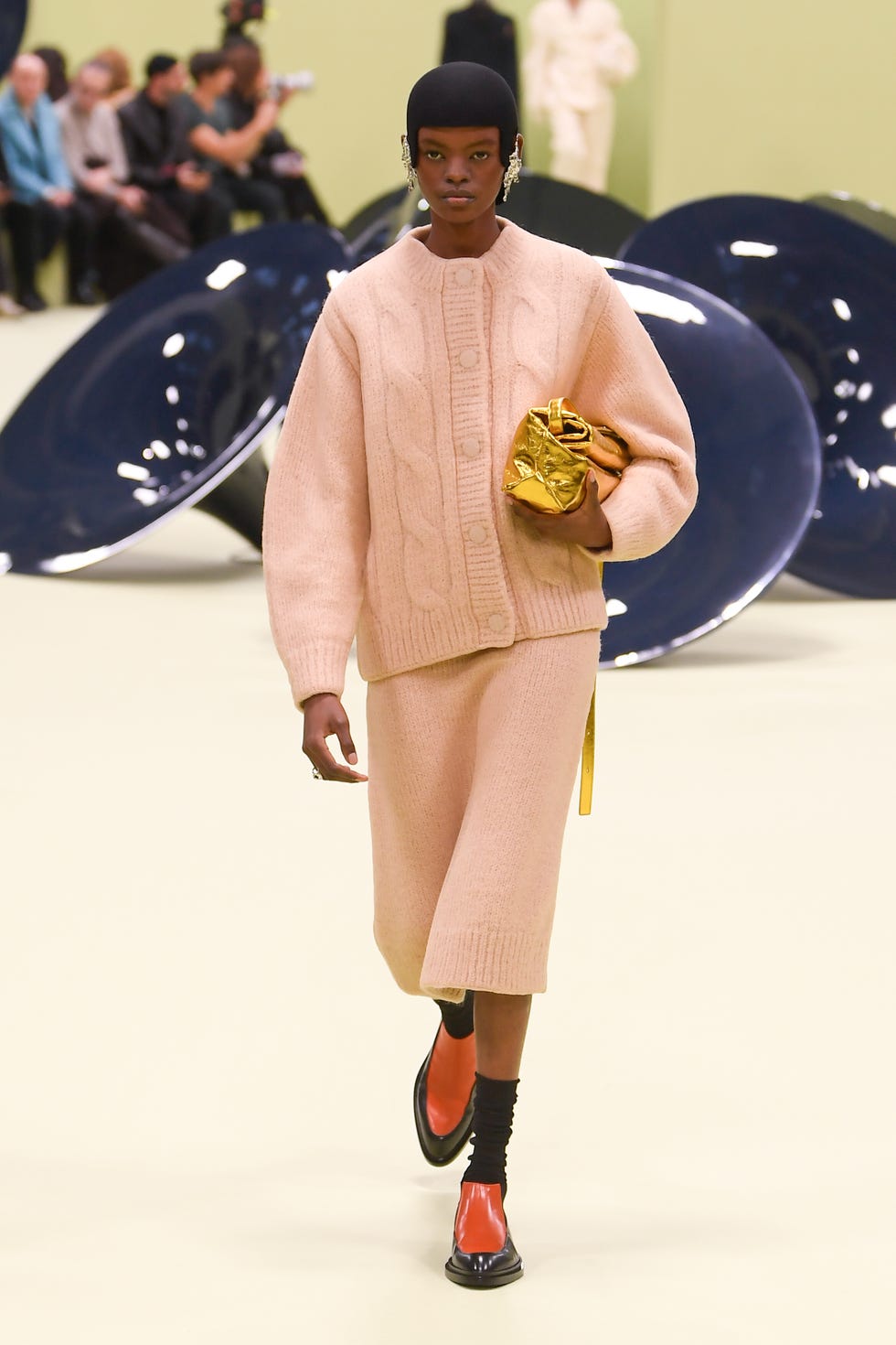 un mannequin portant un cardigan en tricot pêche et une jupe en tricot avec des bottes bicolores un sac doré caché sous son bras des boucles d'oreilles argentées et une casquette noire sur le défilé jil sander rtw automne 2024 dans un tour d'horizon des meilleures tenues de cardigan 2024