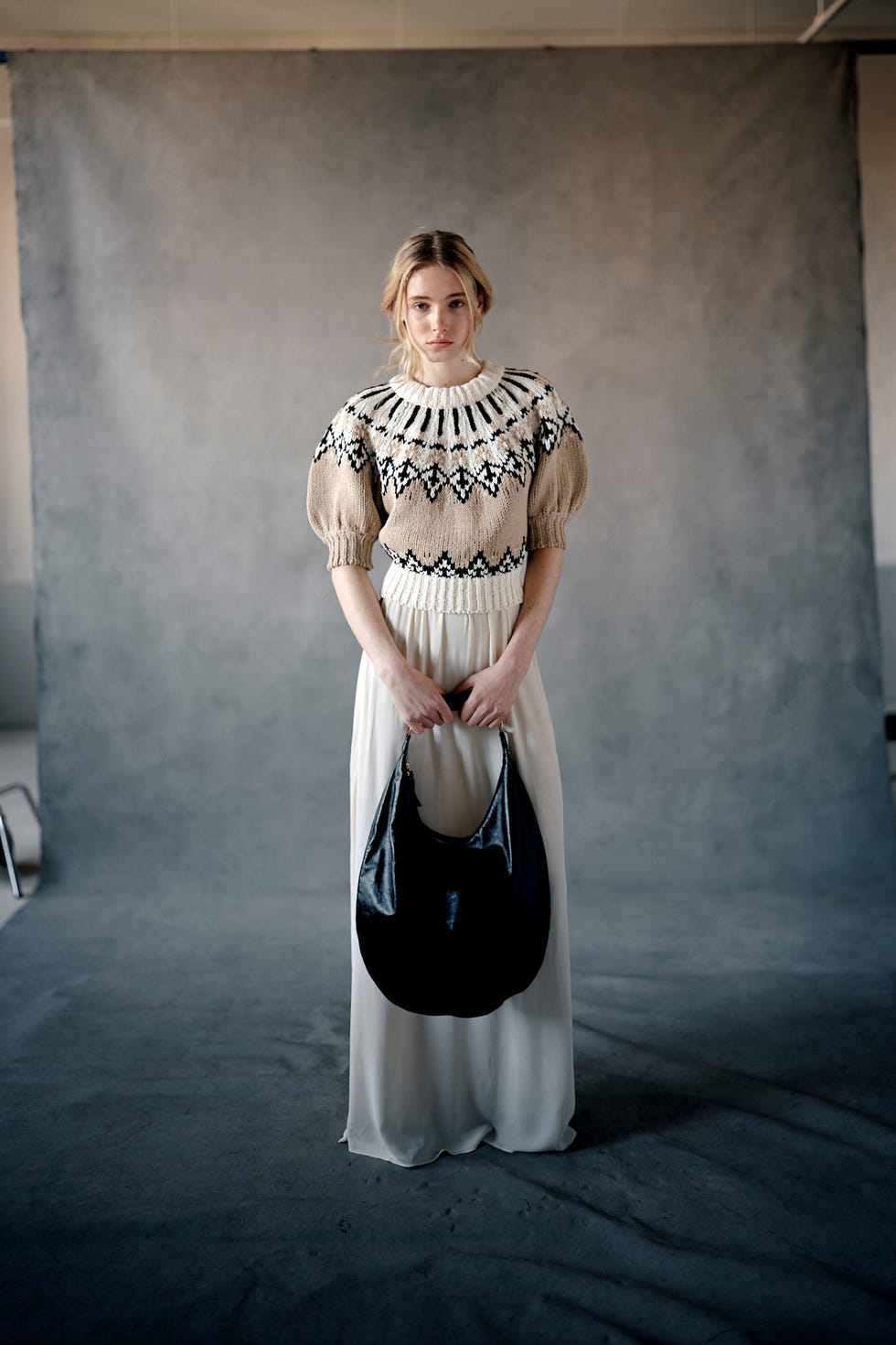 Modèle féminin portant un pull tricoté et une jupe longue tenant un sac à main noir