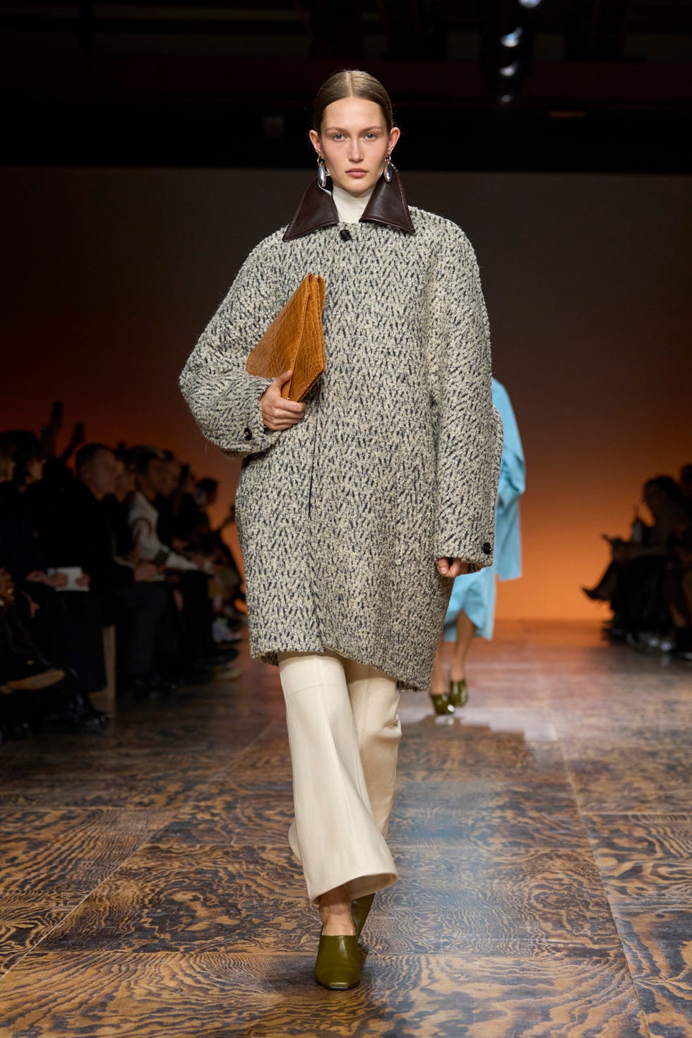 Mannequin marchant sur la piste portant un manteau texturé et des chaussures tenant une pochette