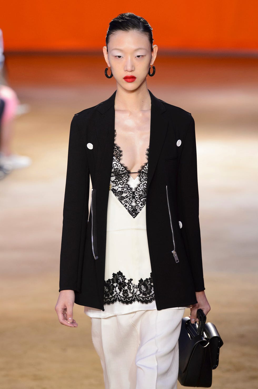 Mannequin portant un élégant blazer noir sur une camisole avec des détails en dentelle et un pantalon de couleur crème tenant un petit sac à main noir