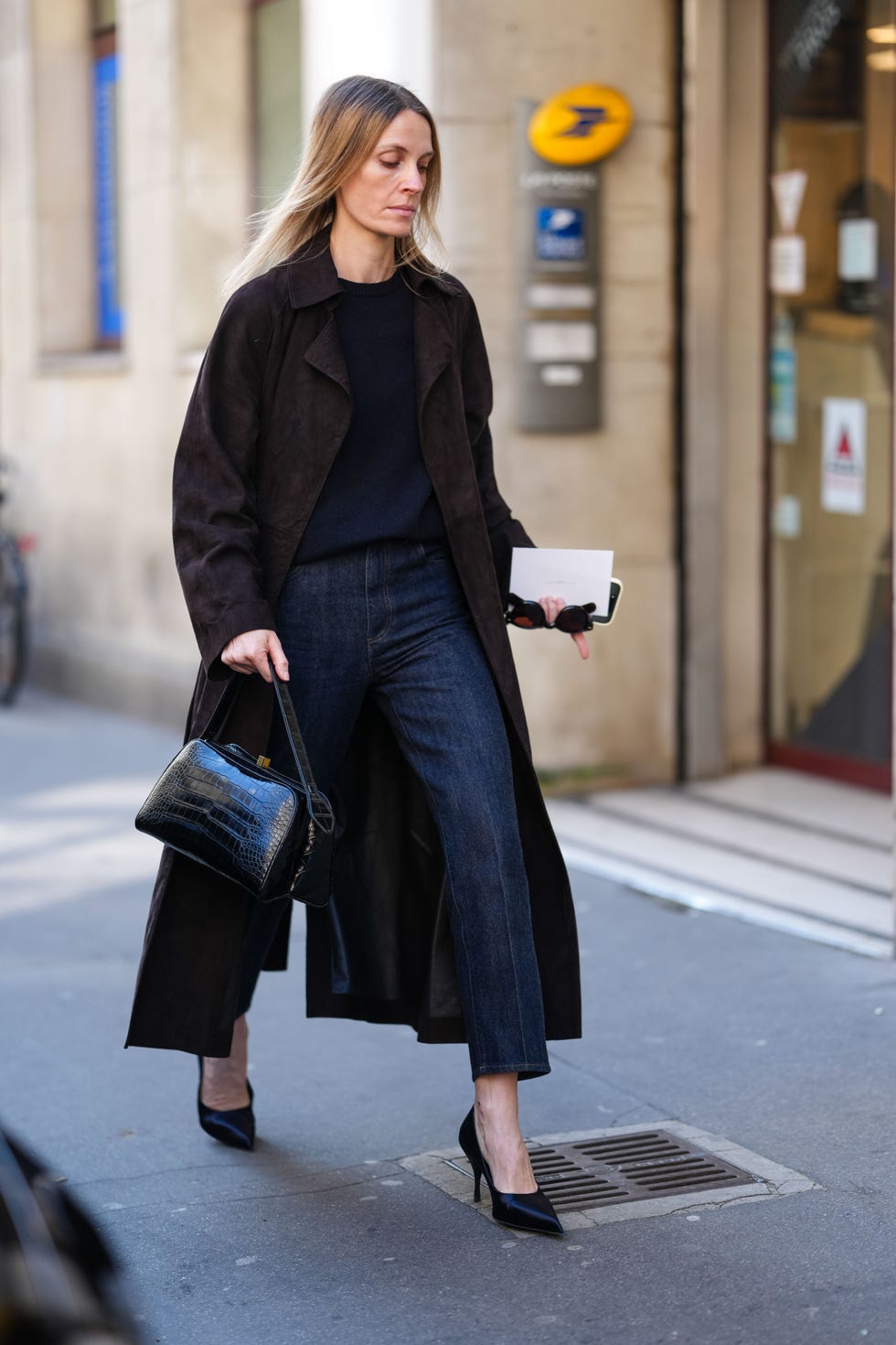 Paris, France 05 mars: Un invité porte un pull noir, un trench en daim brun foncé, un pantalon de jean en jean bleu marine, un sac en cuir en peau de serpent noir brillant, des lunettes de soleil noires à la main, des talons à bout pointu brillant, à l'extérieur de la rangée, pendant la France de la mode Paris, l'automne / hiver 2025 2026 le 5 mars 2025 à Paris, France. (Photo d'Images d'Edward BertheLot / Getty)