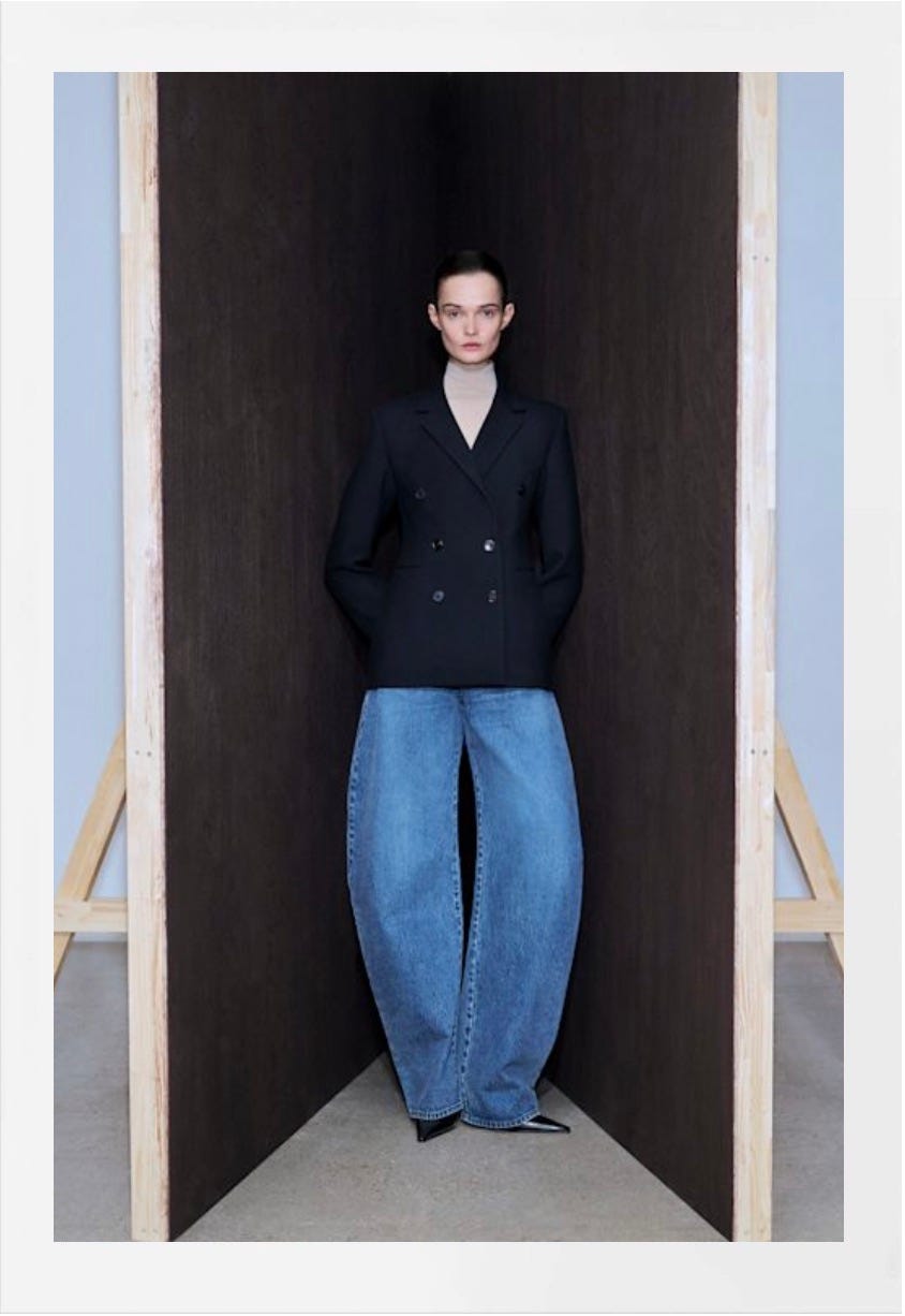 un mannequin se tient entre deux panneaux en bois, portant un blazer noir à double boutonnage et un jean surdimensionné