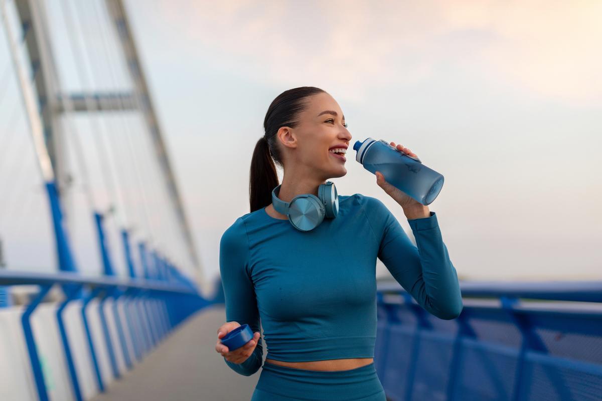 Jeune femme excitée prenant une pause après son jogging, faisant une pause et buvant de l'eau, tenant une bouteille pour se rafraîchir pendant l'entraînement en plein air le soir