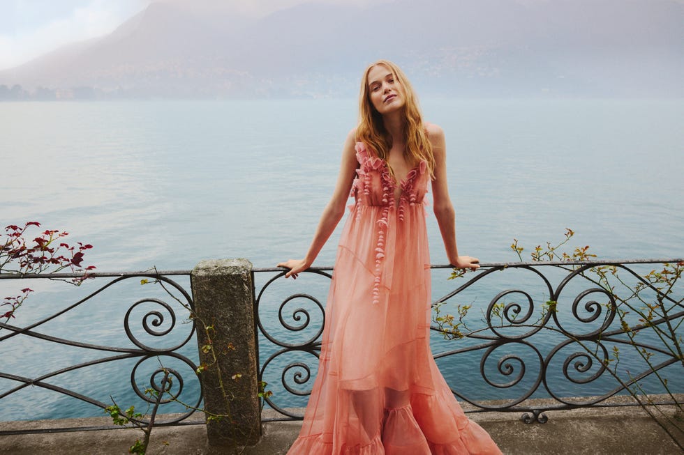 Une silhouette vêtue d’une robe rose fluide debout près d’une balustrade surplombant un plan d’eau.